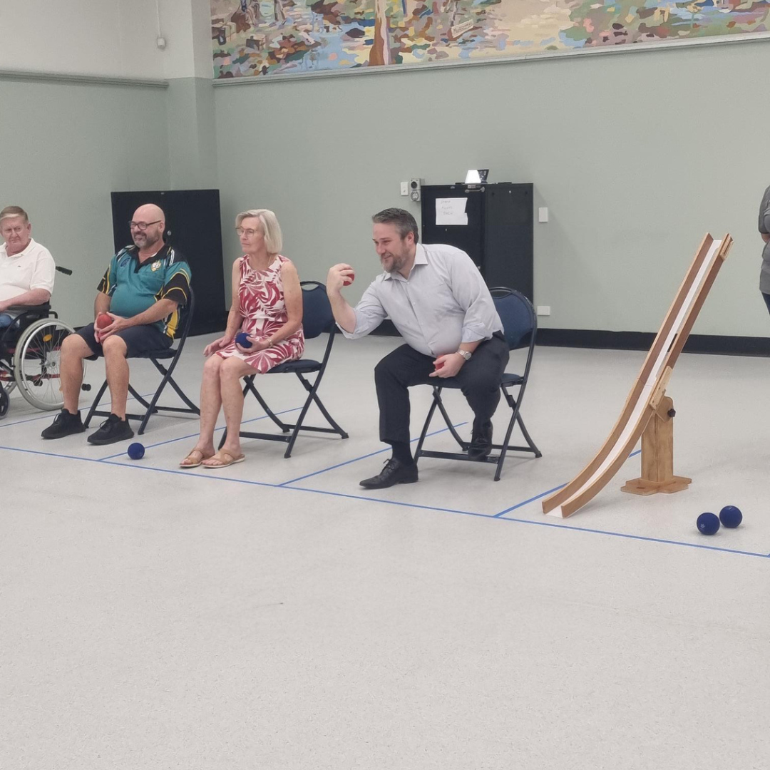 A seated man rolls a small blue ball toward a wooden ramp during an inclusive sport activity.