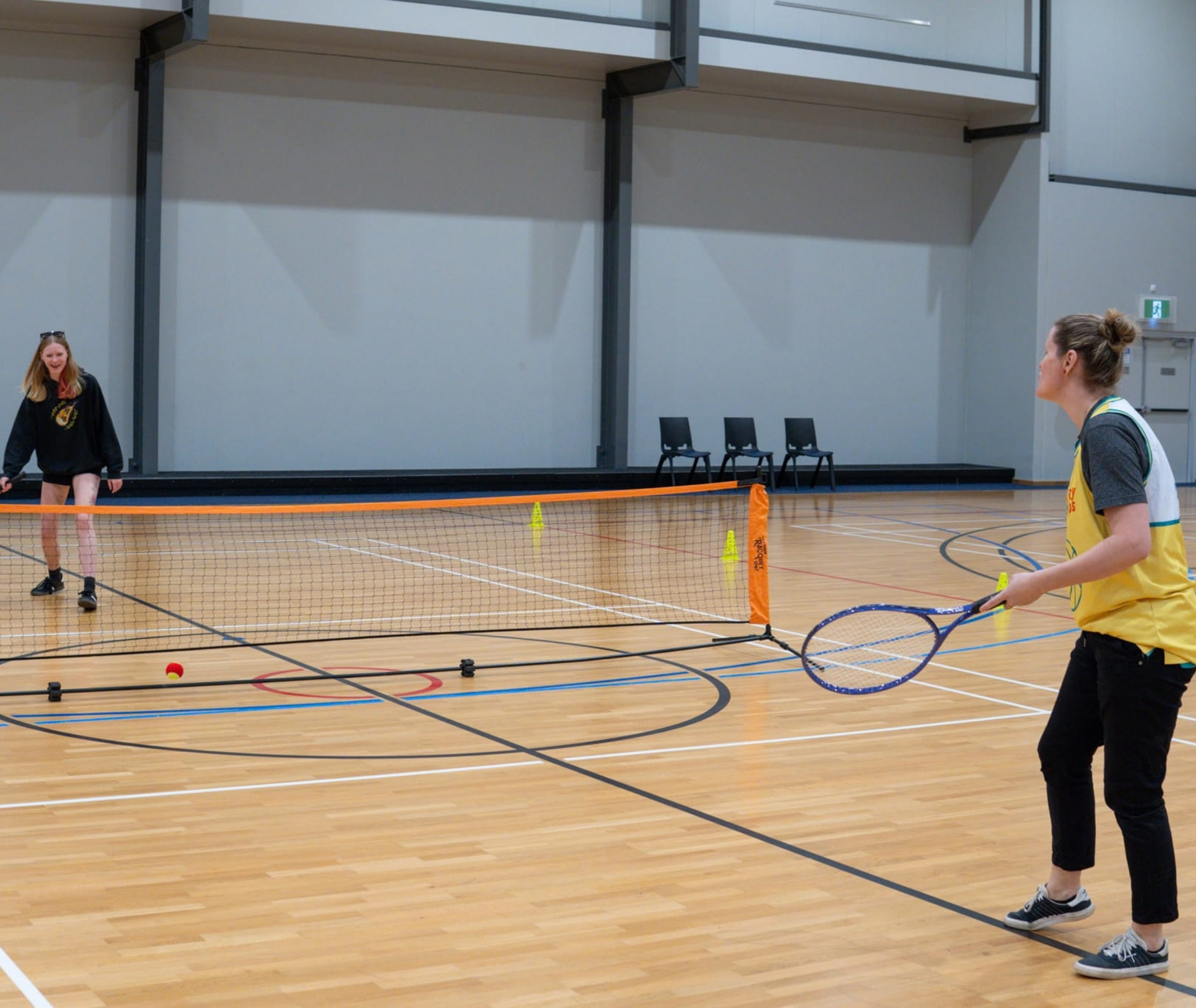 Two people play tennis across a low net on an indoor court using modified equipment.