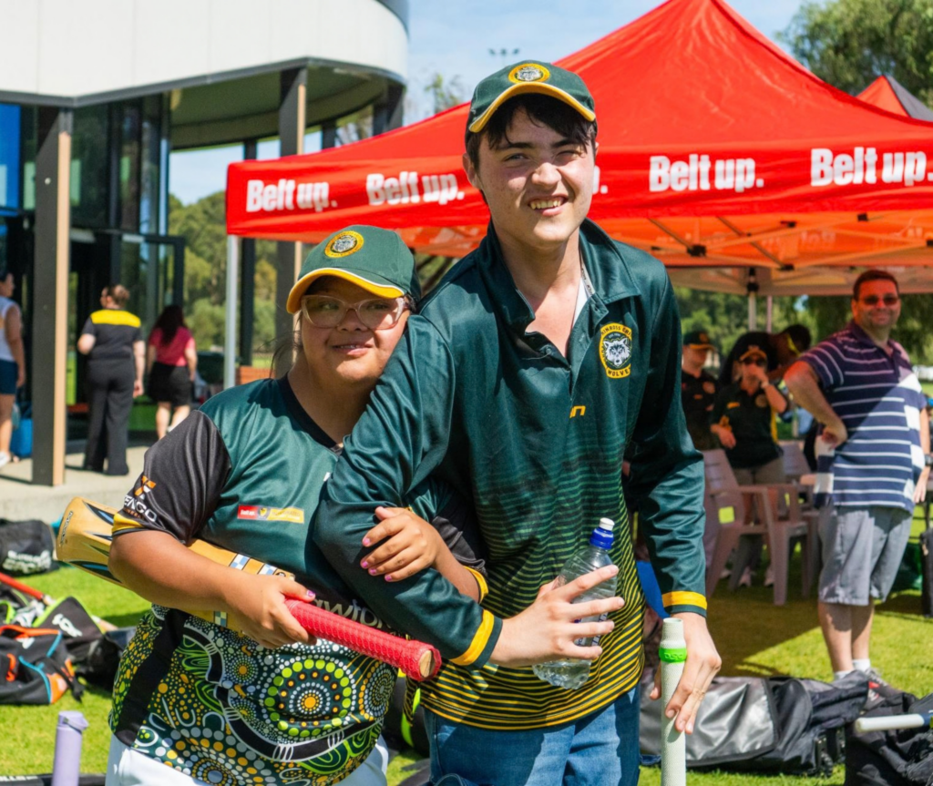 Two young cricketers in green uniforms pose together outdoors, with cricket bats and equipment nearby.