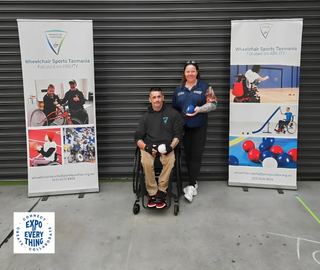 A wheelchair user and Sport4All representative stand with Wheelchair Sports Tasmania banners.