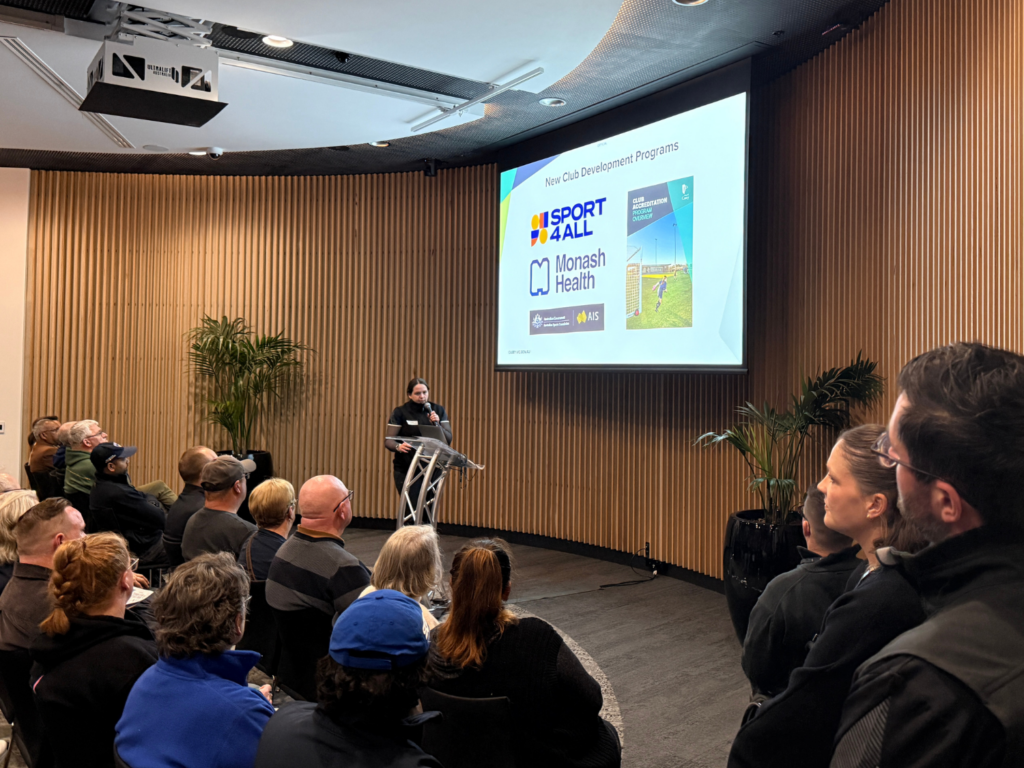 A presenter addresses an audience at a City of Casey club development event, with a slide about Sport4All, Monash Health and the Club Accreditation Program displayed behind them.