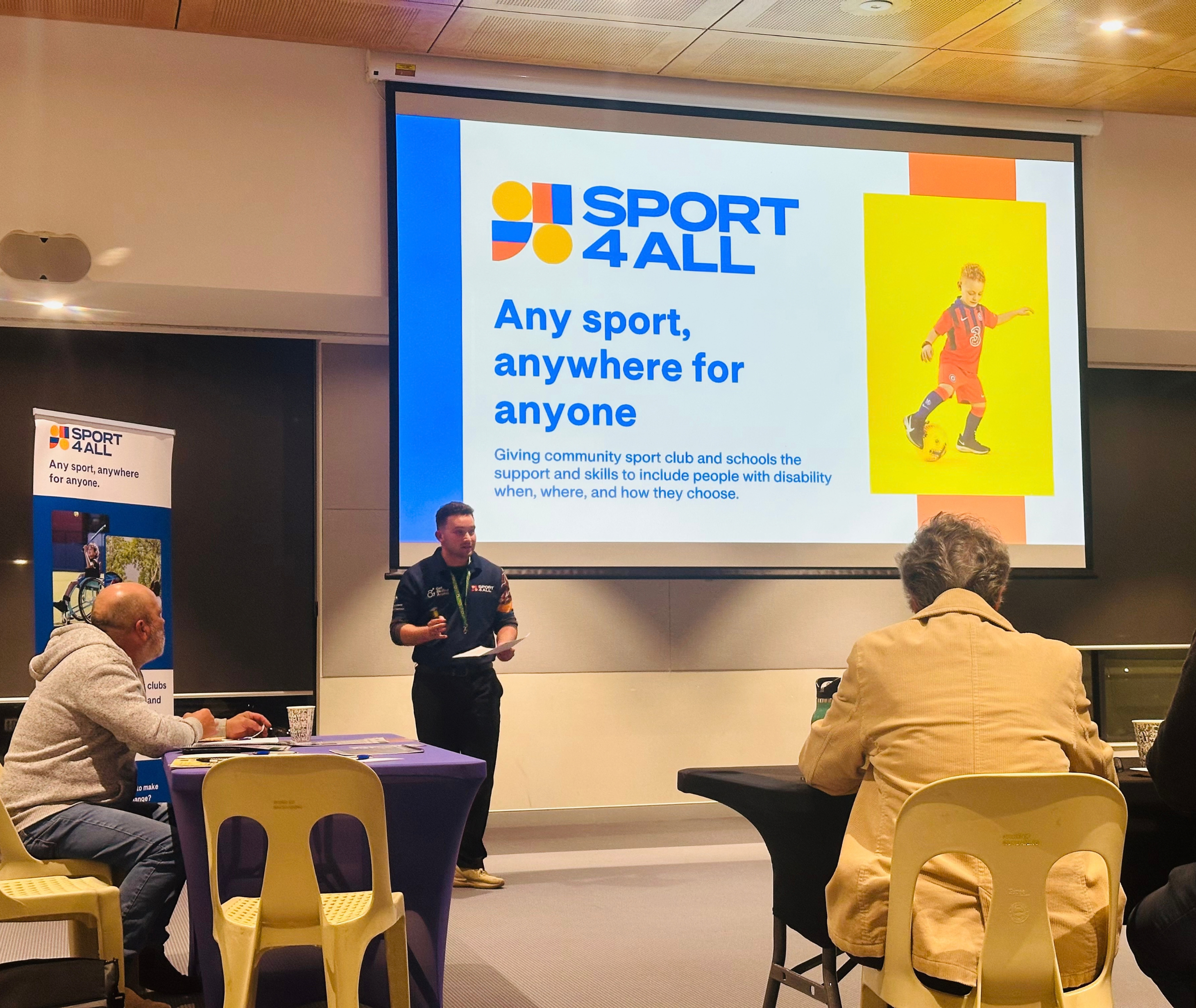 Inclusion Coach, Michael, is speaking in front of the room with Sport4All slides displayed behind him on thr big screen.