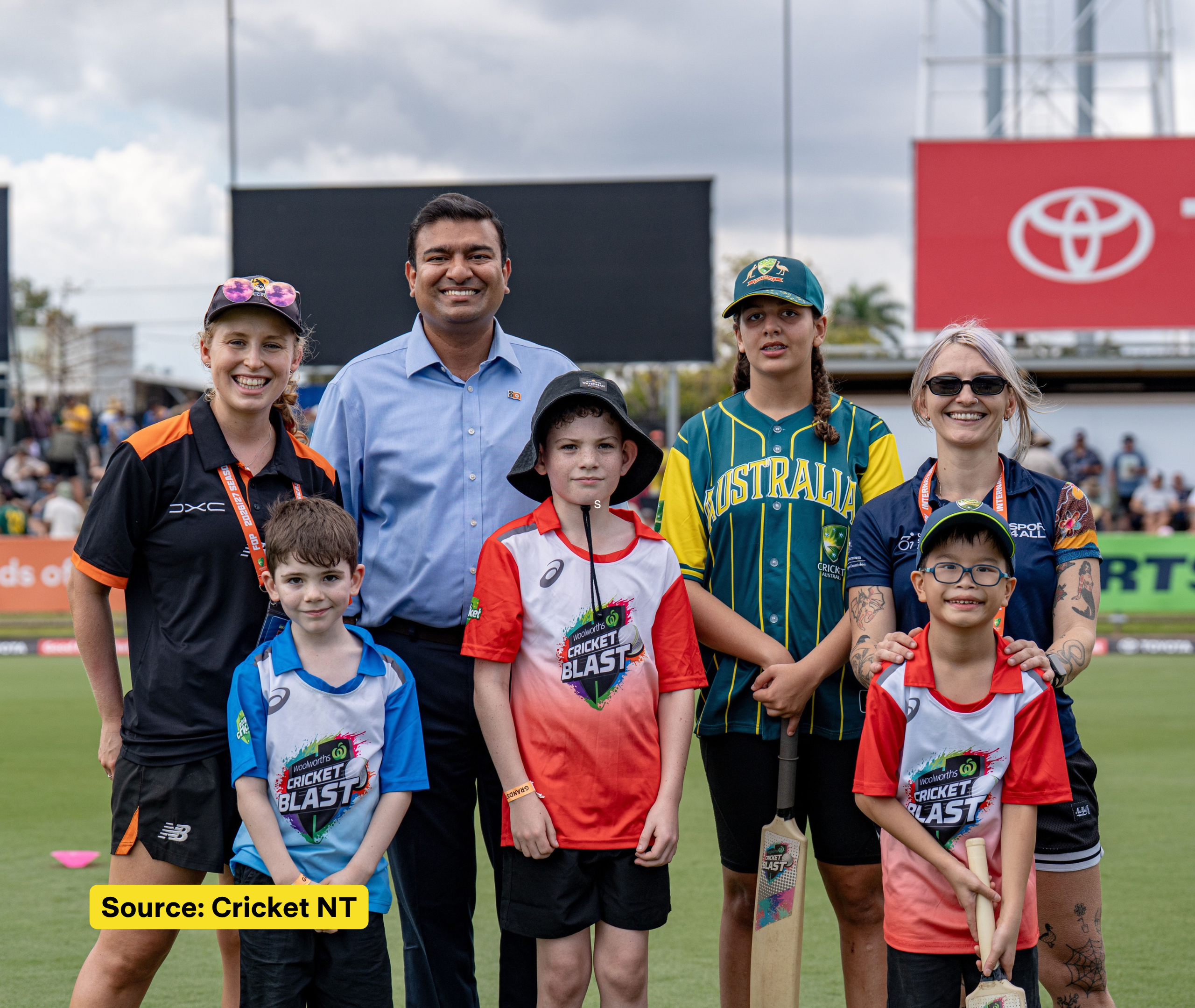 Children in Cricket Blast shirts stand with adults, including a Sport4All team member, for a group photo on a cricket ground.