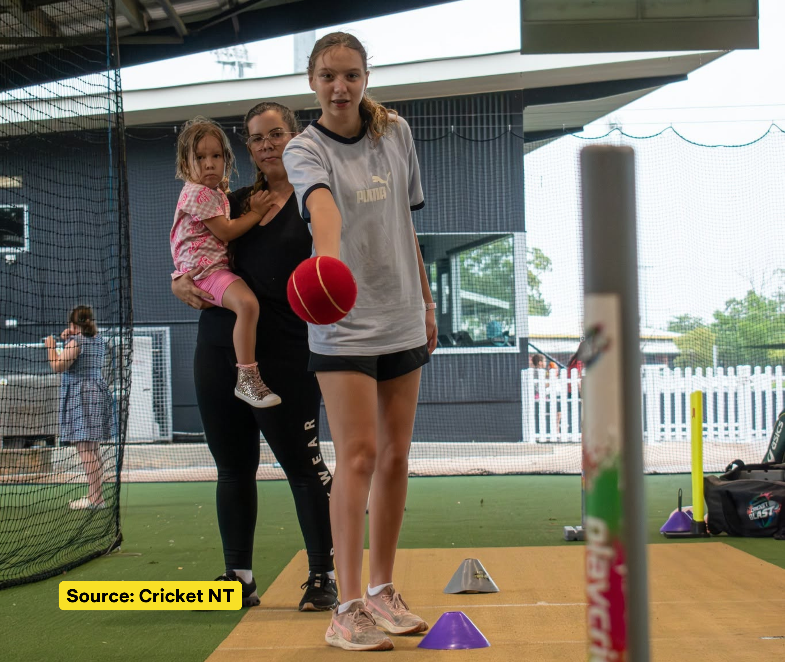 A young participant practises an underarm throw with a red ball inside a cricket net, while an adult holding a child watches behind.