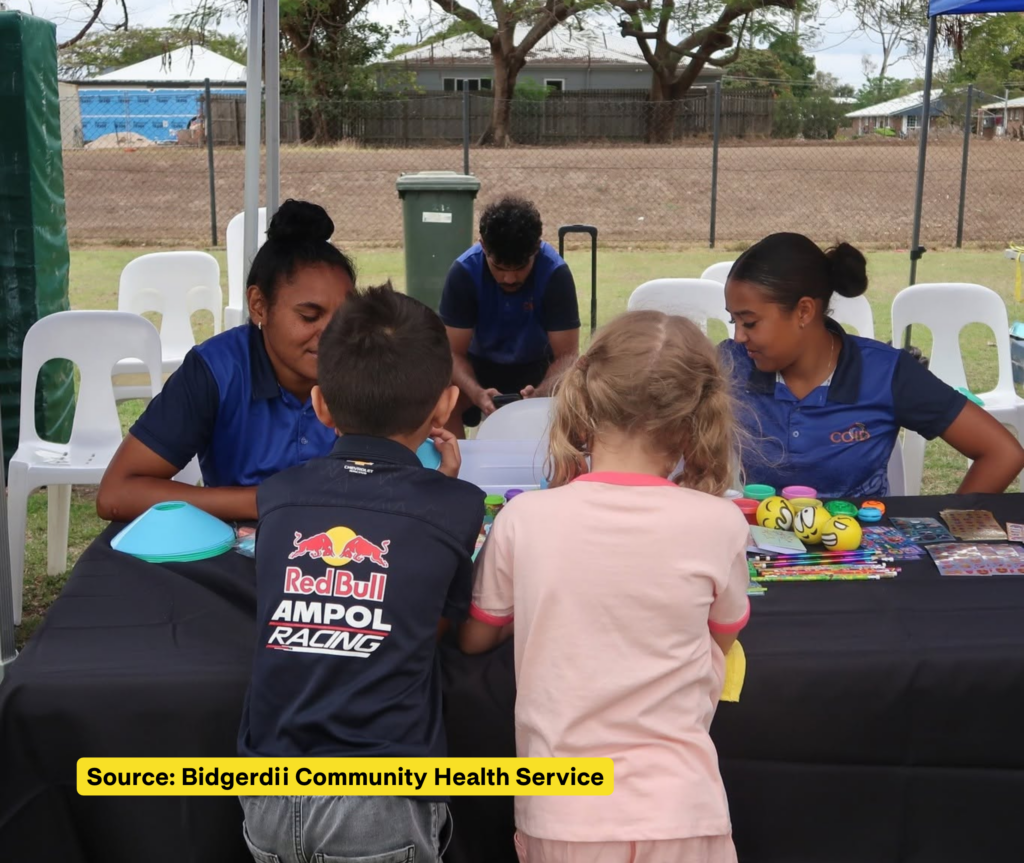 Two children explore activity materials at a table with two staff members, with colourful balls, pencils and stickers spread across the table.
