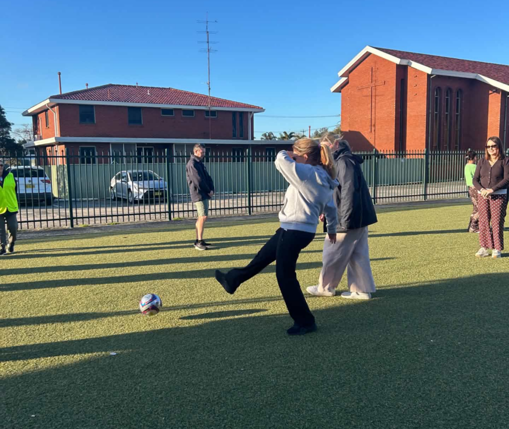 A participant steps towards a soccer ball on an outdoor artificial turf court while other adults watch.